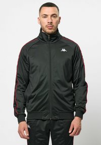 Zwarte sportieve jack met rode zijstrepen, hoge kraag en rits aan de voorkant. Gemaakt van gladde stof, met logo op de borst en geribbelde manchetten.