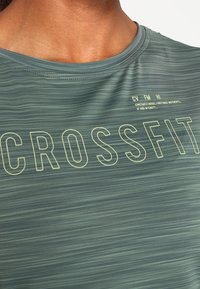 Grön sporttröja med en strukturerad design som har ordet "CROSSFIT" i fet, ljusgul text på framsidan, rund halsringning.