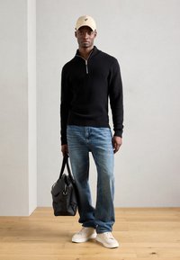 Black knitted sweater with a zip collar, light blue jeans, white sneakers, and a beige cap. Model holds a black leather bag.
