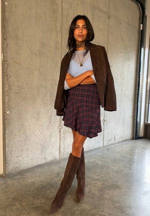 Femme debout, bras croisés, portant une veste marron sur un haut clair, une jupe à carreaux, de grandes bottes marron et une écharpe à motifs autour du cou, contre un mur en béton.