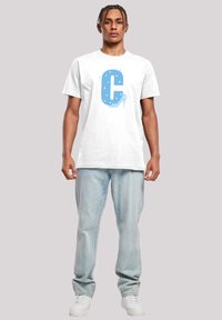 Hvit t-skjorte med korte ermer som har en stor blå bokstav "C" med stjerneakenter og et grafisk motiv av en høyhælte sko, sammen med lyseblå jeans.
