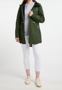 Veste imperméable verte avec une capuche noire, une fermeture éclair à l'avant, deux poches latérales et un logo sur la poitrine, portée par-dessus une chemise grise et un pantalon blanc.