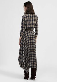 Vestito lungo a quadri con orlo asimmetrico, caratterizzato da motivi a scacchi blu navy e beige. Vita aderente con cintura marrone, maniche lunghe e tessuto morbido.