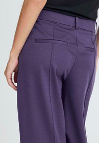 Pantalons violets en tissu texturé, dotés de poches arrière et d'une taille lisse. La coupe est ajustée au niveau des hanches et ample au niveau des jambes.