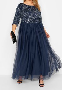 Námořnická modrá večerní šaty s tříčtvrtečními rukávy, korzet zdobený flitry a splývavá tylová sukně. V jedné ruce drží černou kabelku.