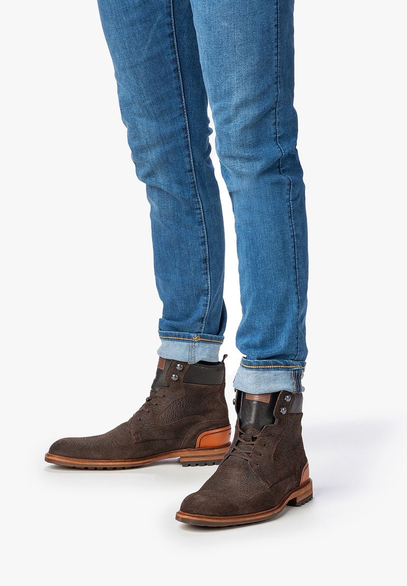 Botas de cuero marrón hasta el tobillo con parte superior texturizada, acento en el tacón de color marrón contrastante y diseño de cordones. Llevadas con jeans azules enrollados.