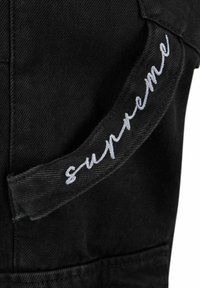 Schwarzer Denim-Stoff mit glänzender Oberfläche, der entlang des Randes des Kleidungsstücks ein gesticktes "Supreme"-Logo in Weiß aufweist.