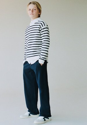 Jeune personne debout, portant un pull à rayures blanches et noires, une chemise blanche, un pantalon ample bleu marine et des baskets blanches, sur un fond uni.