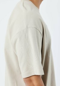 Camiseta de algodón beige con mangas cortas y patrones de hojas bordadas en blanco en la manga izquierda, con cuello redondo y corte relajado.