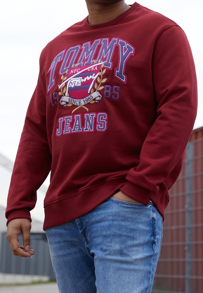 Person trägt ein rotes Tommy Jeans Sweatshirt mit Logo und blaue Jeans, steht im Freien mit einer Hand in der Tasche.