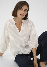 Weiße Bluse mit floraler Stickerei, leichtem Stoff, lockerem Schnitt, Puffärmeln und einem Knopfverschluss am Ausschnitt. Kombiniert mit dunkelblauen Denim-Hosen.