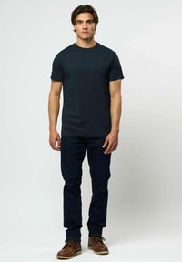 Jauns vīrietis stāv, valkājot vienkāršu tumši zilu T-kreklu, tumšas džinsas un brūnas ādas zābakus uz gaiša fona.