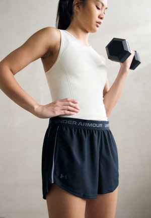 Sorte atletiske shorts med et elastisk Under Armour livbånd, parret med en tætsiddende hvid tanktop og en håndvægt i den ene hånd.