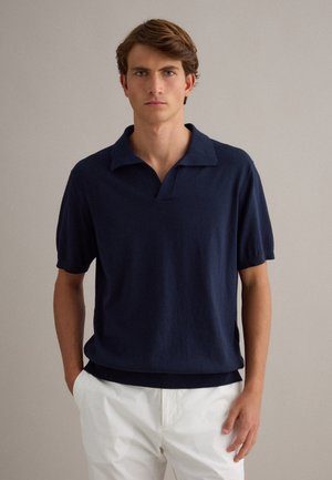 Hombre joven con polo azul marino de manga corta y pantalones blancos, de pie con las manos en los bolsillos contra un fondo liso.