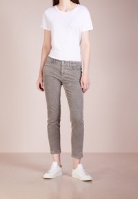 Bílý tričko s krátkým rukávem spárované se světle šedými slim-fit manšestrovými kalhotami a bílými teniskami na jednobarevném pastelovém pozadí.