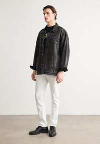 Zwarte denimjas met knoopsluiting, twee voorzakken, gedragen over een zwart shirt en gecombineerd met witte broek en zwarte schoenen.