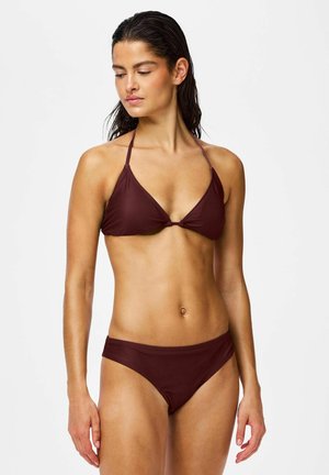 Femeie cu păr închis la culoare ud, purtând un bikini roșu închis, privind în jos și spre o parte pe un fundal simplu și deschis.