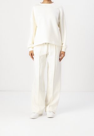 Osoba ubrana w luźną białą bluzkę z długim rękawem, szerokie białe spodnie i białe trampki, stojąca na jednolitym białym tle.