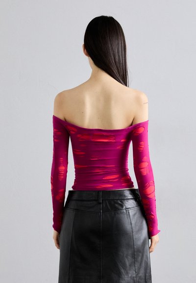 Fitted, off-the-shoulder top in vibrant pink with red, textural cutouts. Long sleeves. Paired with a high-waisted black leather skirt.