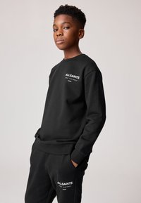 Mladý člověk stojí s rukama v kapse, oblečený v černé mikině AllSaints a odpovídajících černých kalhotách na jednobarevném světlém pozadí.