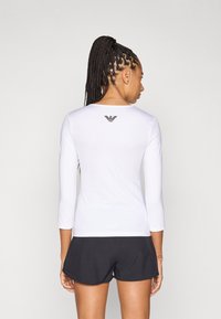 Top desportivo branco de manga comprida com gola redonda e detalhe de logótipo preto nas costas, combinado com calções desportivos pretos. Textura suave.