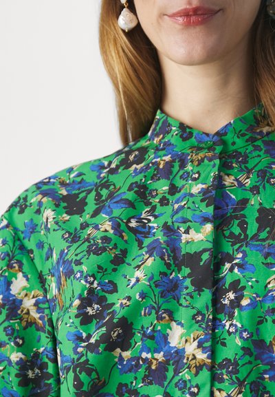 Camicia floreale verde con fiori blu e neri, colletto alla coreana e chiusura a bottoni, realizzata in un tessuto leggero con una texture liscia.
