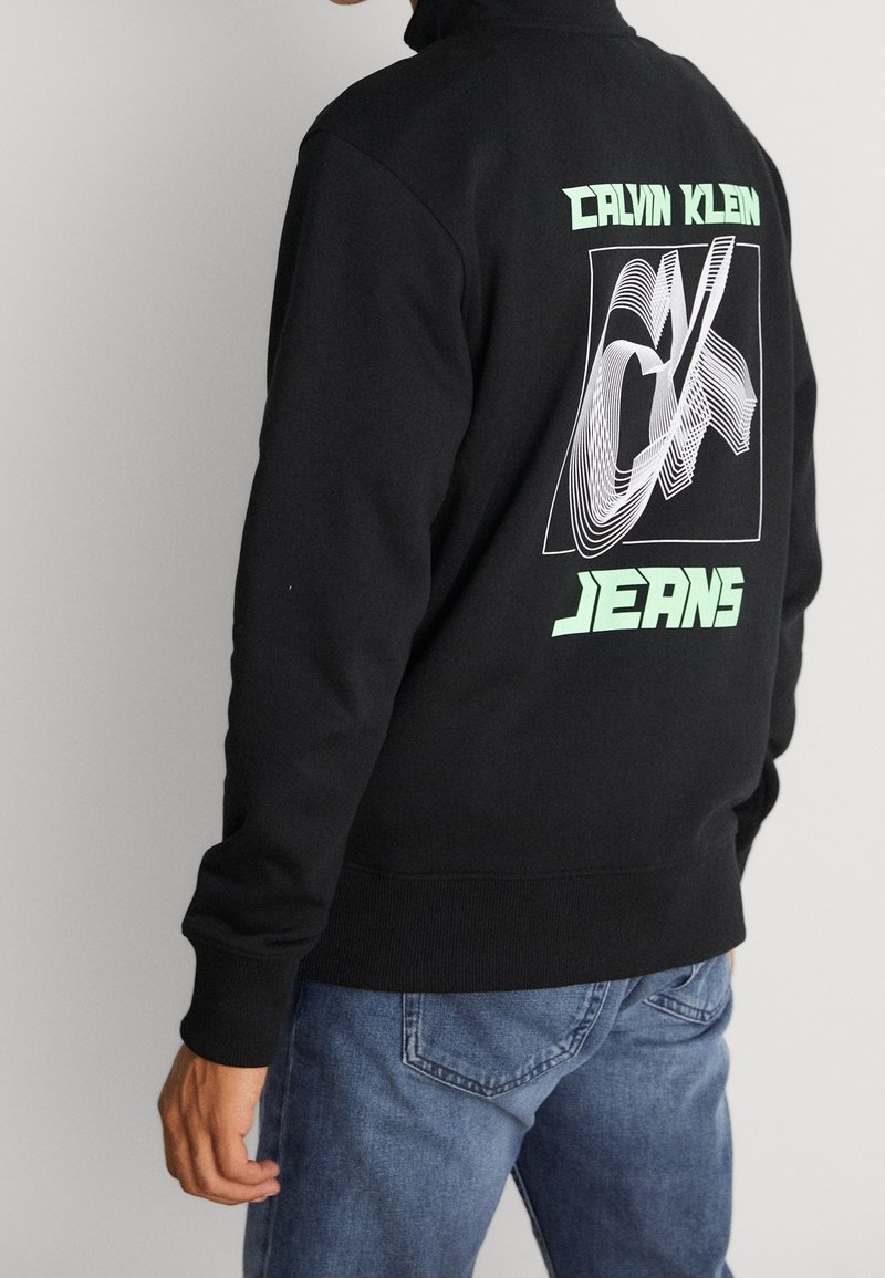 Personne portant un sweat-shirt Calvin Klein Jeans noir avec un logo vert néon et blanc au dos, associé à un jean bleu.