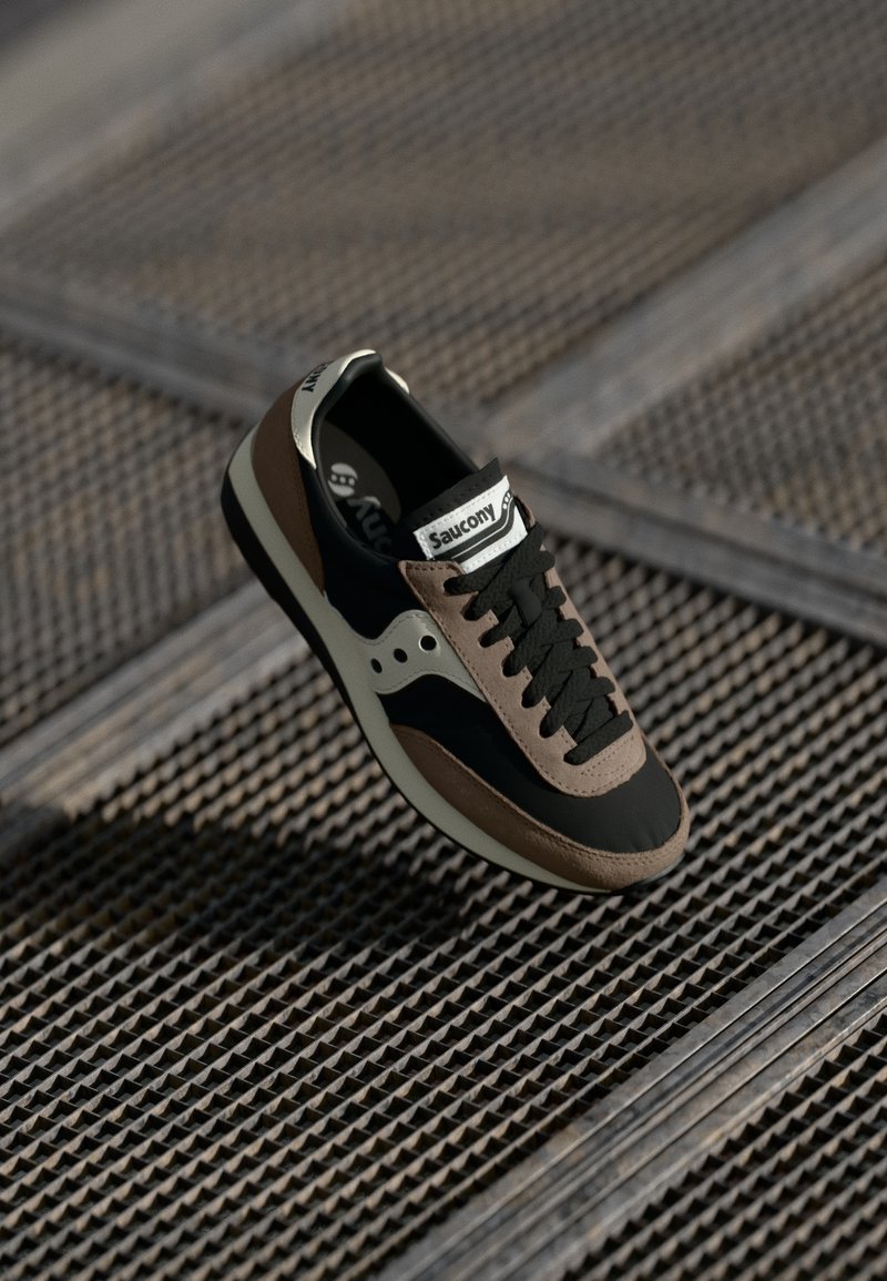 Melnas un brūnas Saucony sporta kedas ar melnām kurpēm uz metāla režģa virsmas, apgaismotas ar dabīgo gaismu, kas rada ēnas.