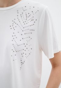 Camiseta blanca de manga corta con un patrón de tachuelas plateadas dispuestas en un diseño gráfico en la parte frontal. Tela suave con textura lisa.