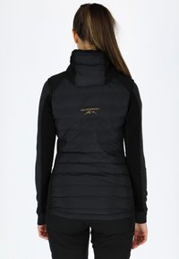 Gilet imbottito nero con texture trapuntata e colletto alto, abbinato a maniche lunghe scure. Presenta un logo dorato sulla parte superiore della schiena.