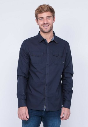 Chemise à manches longues bleu marine avec deux poches sur la poitrine, col contrasté et coupe droite. Portée avec un jean en denim.