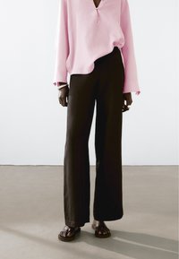 Dlouhé černé kalhoty volného střihu v kombinaci s světle růžovou košilí na knoflíky s uvolněným výstřihem. Oblečení doplňují hnědé sandály.