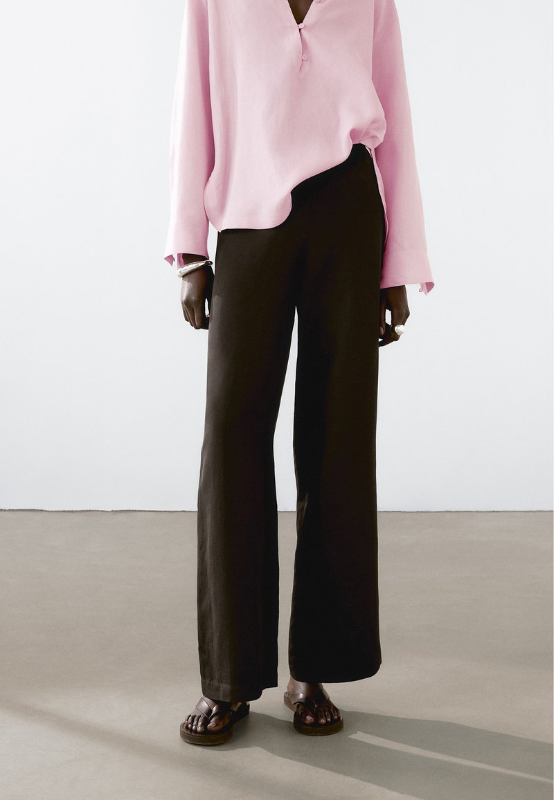 Dlouhé černé kalhoty volného střihu v kombinaci s světle růžovou košilí na knoflíky s uvolněným výst�řihem. Oblečení doplňují hnědé sandály.