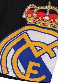 Czarne szorty z logo Real Madryt. Logo przedstawia żółtą koronę nad niebieskimi i białymi literami, z wyraźnymi, zakrzywionymi kształtami.