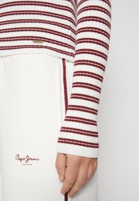 Maglione bianco a costine con strisce bordeaux, caratterizzato da un design corto e un'etichetta con logo. Abbinato a pantaloni bianchi con un accento bordeaux.