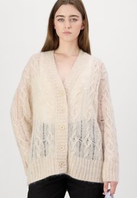 Cardigan tricotat voluminos în bej deschis, cu modele cu cabluri, decolteu în V, cinci butoane și tiv ribbed. Textură moale cu secțiuni transparente.