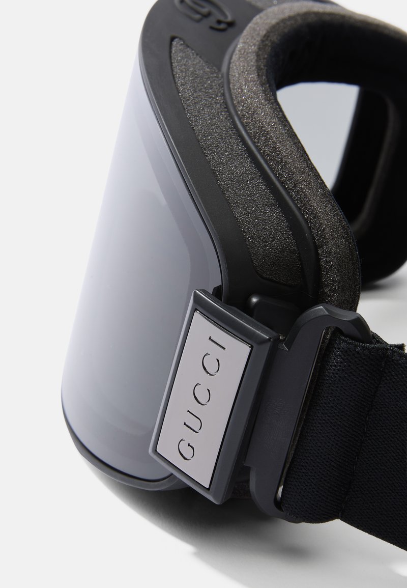 Ski goggles with a clear, mirrored lens, black foam padding, and adjustable black strap featuring a silver logo plate. Smooth, modern design.