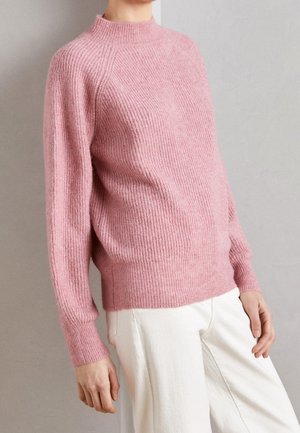 Personne portant un pull en maille côtelée rose à manches longues associé à un pantalon blanc, debout devant un fond gris uni.