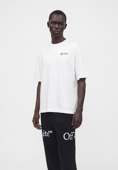Homme portant un t-shirt blanc Off-White et un pantalon noir avec de grands logos Off-White, debout devant un fond clair uni.