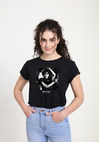 Woman with curly hair wearing black T-shirt featuring black-and-white image of a woman dancing, paired with light blue jeans, hands in pockets.