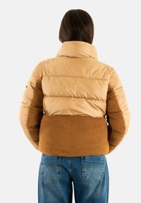 Vadderad jacka med en tanfärgad glansig övre sektion och en brun ludden nedre sektion. Har en hög krage och en vid passform, kombinerad med blå jeans.