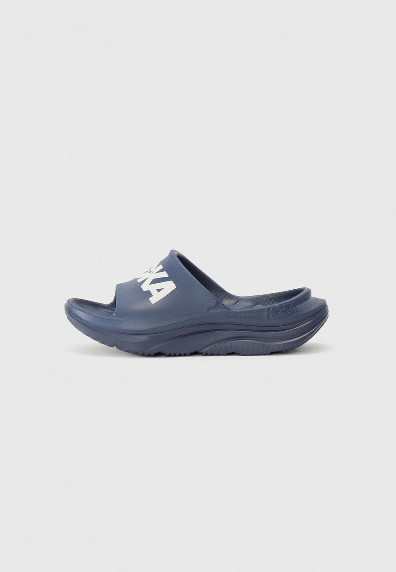 Jūras zila slīdėjsandaļu pāris, kas izgatavots no gumijas ar gludu tekstūru. Iezīmējas ar atvērtu pirkstu daļu, plašu siksnu un izteiksmīgu baltu logotipu sānos.