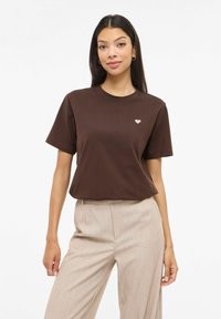 Maglietta di cotone marrone con maniche corte, caratterizzata da una piccola grafica a cuore chiaro sul petto a sinistra. Abbinata a pantaloni beige chiaro.