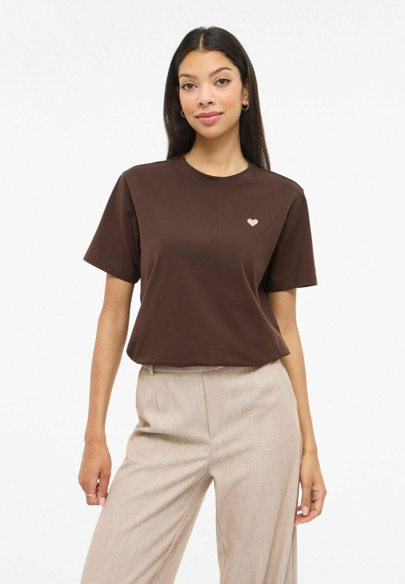 Maglietta di cotone marrone con maniche corte, caratterizzata da una piccola grafica a cuore chiaro sul petto a sinistra. Abbinata a pantaloni beige chiaro.