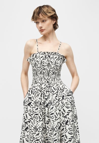 Γυναίκα που φοράει μαύρο και άσπρο φόρεμα με floral σχέδιο, χωρίς μανίκια και λεπτές τιράντες, με τα χέρια στις τσέπες, κοιτάζοντας ελαφρώς στο πλάι.