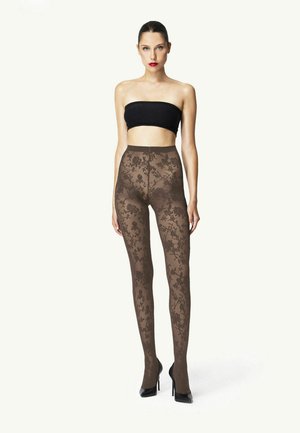 Frau trägt schwarzen trägerlosen Oberteil, transparente braune Strumpfhose mit Blumenmuster und schwarze High Heels, steht vor hellem einfarbigem Hintergrund.