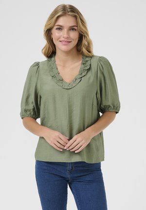 Blouse verte avec un décolleté en V froncé, manches courtes à volants et une texture douce. Associée à un jean en denim bleu, design minimaliste et décontracté.