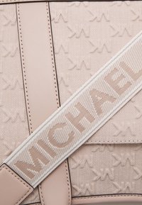 Beżowa torba na ramię z tkaniny, z skórzanymi wykończeniami, posiadająca szeroki, teksturowany pasek na ramię wyszyty nazwą marki "NICHARL."