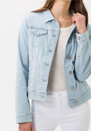 Vrouw draagt een lichtblauwe spijkerjas over een wit shirt en witte broek, met één hand die de kraag van het jasje aanpast.
