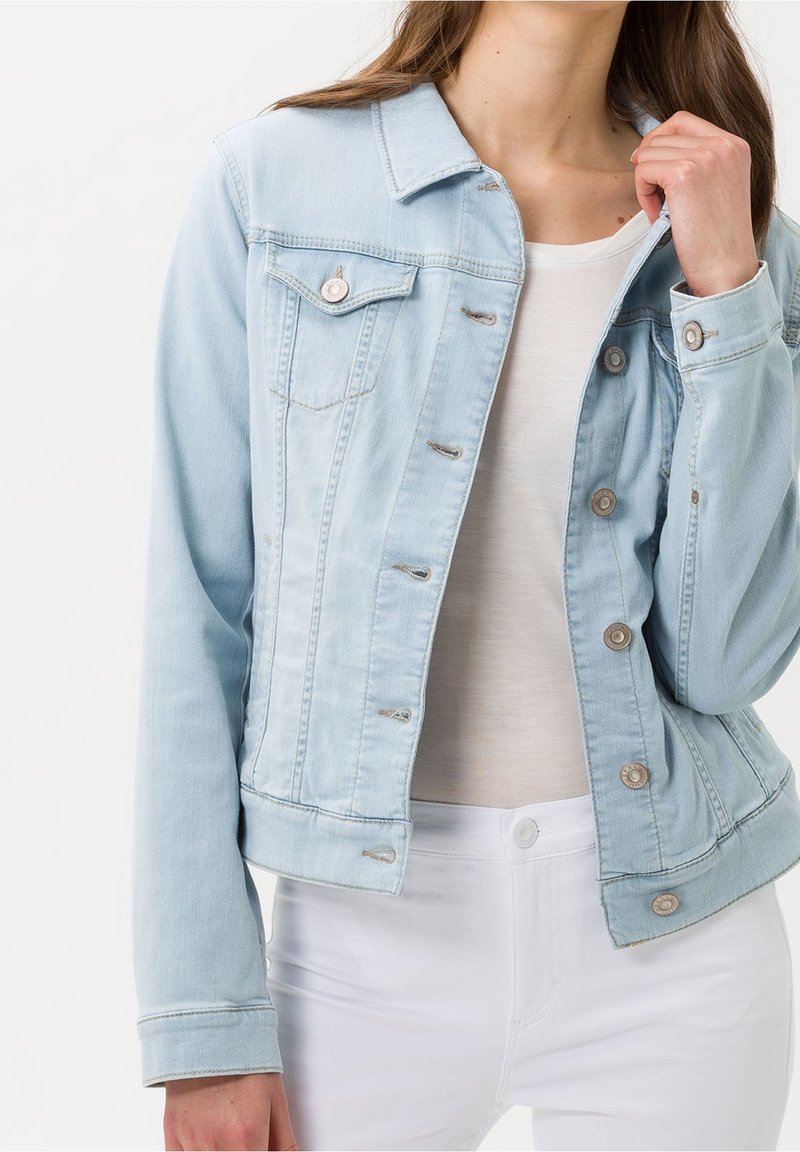 Femme portant une veste en jean bleu clair sur un haut blanc et un pantalon blanc, ajustant le col de la veste d'une main.
